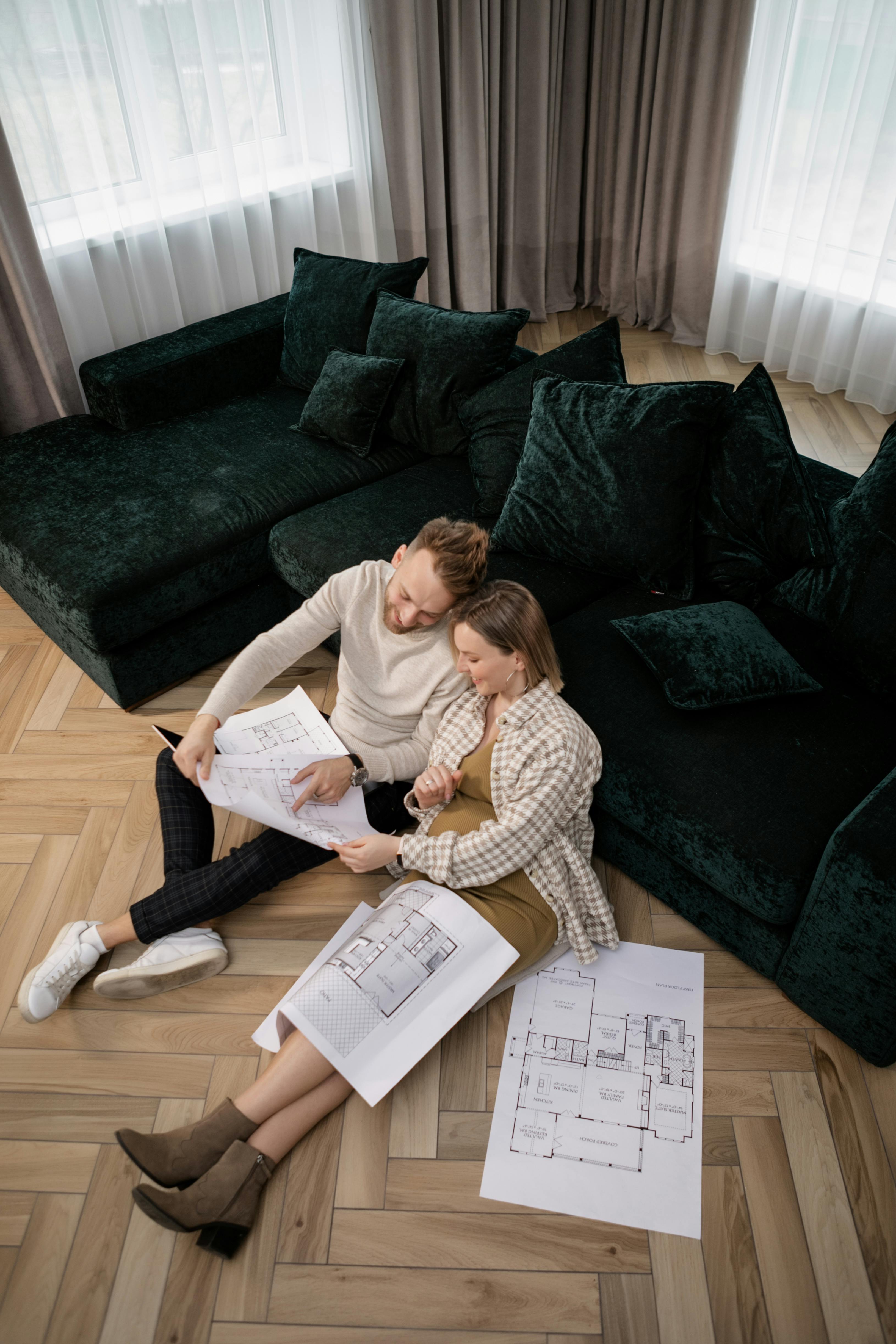 A young couple sits on the floor reviewing house blueprints in a modern living room setting.
